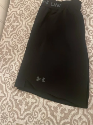 Svarta träningstights Under Armour - Säljer ett par svarta träningstights från Under Armour med logga på benet och bred resår i midjan med texten 'Under Armour'. Tillverkade i stretchigt funktionsmaterial som passar perfekt till gymmet eller löpning.