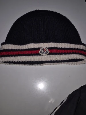 Svart Moncler mössa med ränder - Snygg svart ribbstickad mössa från Moncler med vit och röd rand runt kanten samt Moncler-logga framtill. Klassisk design. STORLEK M. Moncler