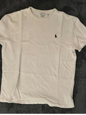 Vit t-shirt från Polo Ralph Lauren - Klassisk vit t-shirt från Polo Ralph Lauren med korta ärmar och rund hals. Liten broderad logga på bröstet i mörkblått. Perfekt basplagg i mjuk bomull som passar till allt.