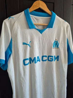 Marseille matchtröja Puma  - Säljer en vit och ljusblå matchtröja från Puma för Olympique de Marseille. Tröjan har klubbmärke och Puma-logga på bröstet, blå detaljer på krage och ärmar samt sponsortryck på magen. Tillverkad i lätt och ventilerande material.