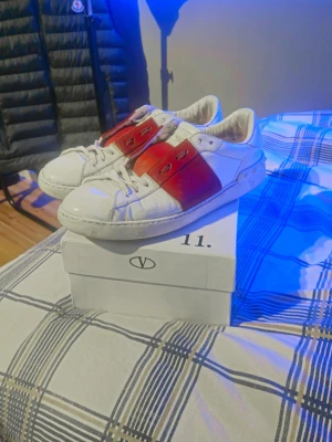 Valentino garavani - Säljer ett par snygga vita sneakers från Valentino Garavani med bred röd läderdetalj över mitten. Klassisk låg modell med snörning och stilren design. Perfekta för dig som vill ha något exklusivt och trendigt i garderoben. Priset går att diskuteras