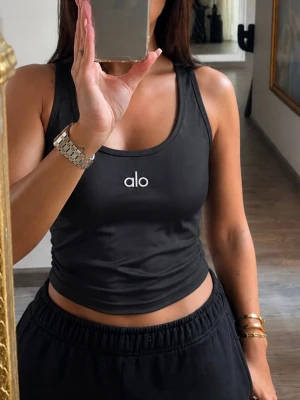 Svart croppad tanktop från Alo - Snygg svart croppad tanktop från Alo med broderad logga framtill. Toppen har en tight passform och är perfekt för en sportig och trendig look. Tillverkad i mjukt och stretchigt material som sitter skönt mot kroppen.