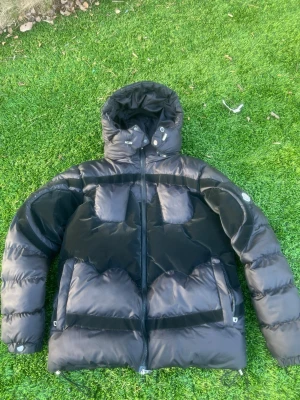 Moncler x Batman puffer jacka - Snygg svart dunjacka från Moncler med klassisk logotyp på ärmen och coolt serietidningsfoder på insidan. Jackan har en puffig design och är tillverkad i glansigt material som ger en exklusiv känsla. Perfekt för kalla vinterdagar. Byte kan också diskuteras 