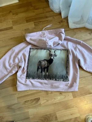 Ljusrosa hoodie med djurmotiv - Unik ljusrosa hoodie med stor patch på ryggen som visar ett konstnärligt tryck av en antilop i naturen. Hoodien har en klassisk huva och är croppad i modellen. Perfekt för dig som vill sticka ut med ett coolt djurmotiv. Materialet känns mjukt och bekvämt.