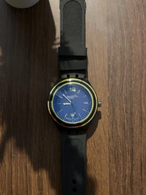 Klocka  - Snygg klocka med blå urtavla, gulddetaljer runt boetten och svart armband i syntetmaterial. Klockan har tydliga markeringar och datumvisning. Perfekt accessoar för dig som gillar stilrena detaljer.