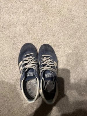Adidas Spezial blå sneakers - Säljer ett par Adidas Spezial sneakers i blå mocka med vita detaljer och klassiska tre ränder på sidan. Skorna har vita snören, svart sula och guldfärgad Spezial-text på sidan. Perfekta för dig som gillar retrostil och streetwear.
