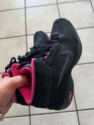 Svarta och rosa Nike high top sneakers storlek 37,5 - Snygga svarta high top sneakers från Nike med rosa detaljer och rosa insida. Skorna har klassisk Nike-logga på sidan, svarta skosnören och en robust sula med AIR-dämpning. Perfekta för dig som vill sticka ut med en sportig och färgstark look.