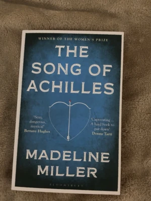 The Song of Achilles av Madeline Miller - En episk roman på engelska av Madeline Miller. Omslaget är blått med vit text och ett hjärta med en pil i mitten. Boken är en modern tolkning av den grekiska myten om Akilles och Patroklos, fylld av kärlek, äventyr och antik dramatik.