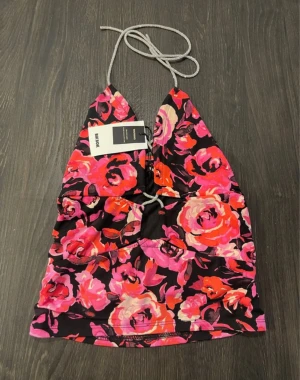 Blommig halterneck backless  topp with crystal tie  - Snygg halterneck topp med blommigt mönster i rosa, rött och beige på svart botten. Toppen har smala knytband i nacken och en liten öppning framtill. Perfekt för dig som vill sticka ut med färg och mönster. Lappen kvar ny pris 279