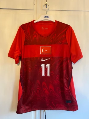 Turkiet fotbollströja Nike röd - Säljer en röd fotbollströja från Nike med Turkiets flagga på bröstet och nummer 11 både fram och bak. På ryggen står det 'Yildiz' och 'Türkiye'. Tröjan har korta ärmar, V-ringning och är tillverkad i ett lätt, andningsbart material perfekt för träning eller match.