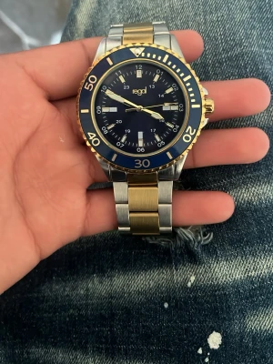 Regal klocka i guld och silver - Snygg klocka från Regal med blå urtavla, guldfärgade detaljer och silverfärgad länk. Klockan har en sportig look med tydliga siffror och markeringar på boetten. Armbandet är i metall med guldfärgade mittlänkar och silverfärgade kanter.