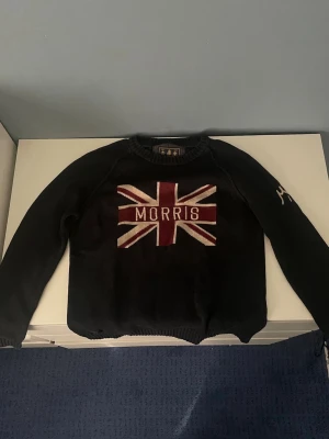 Svart Morris tröja med Union Jack - Snygg svart tröja från Morris med Union Jack-flagga och MORRIS-text broderat på bröstet. Tröjan har rund hals, långa ärmar och ribbade muddar. Perfekt för dig som gillar klassisk brittisk stil och vill sticka ut lite extra.