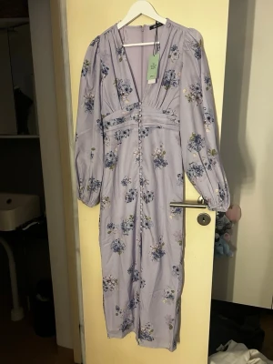 Ljuslila blommig långklänning - Romantisk långklänning i ljuslila med blommigt mönster och v-ringning. Klänningen har långa puffärmar, knappar framtill och markerad midja. Perfekt för sommarens alla tillfällen. Ny med prislapp, slutsålt på hemsidan.