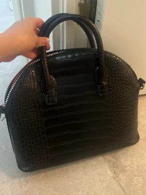 Svart handväska i krokopräglat material - Snygg svart handväska med krokopräglat mönster och rundad form. Väskan har två stadiga handtag och dragkedja upptill. Perfekt för dig som gillar en stilren och trendig look. Använd mycket men i bra skick förutom lite slitningar / repor nere vid kanterna och på botten, men inget som syns direkt när man har på sig den. Öppen för prisförslag