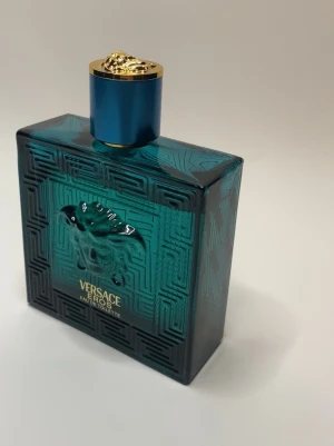 Versace Eros Eau de Toilette - Versace Eros Eau de Toilette i en stilren, turkos glasflaska med guldiga detaljer och ett Medusa-huvud i relief på framsidan. Flaskan har en fyrkantig form och ett blått lock med guldfärgad topp. Perfekt för dig som gillar exklusiva och ikoniska dofter.TESTAD ETT PAR GÅNGER. +BOX INGÅR