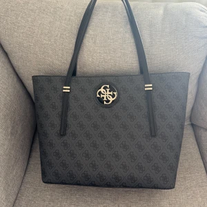 Svart axelväska från Guess med monogram - Snygg svart axelväska från Guess med klassiskt monogrammönster och guldfärgad logga framtill. Väskan har långa handtag, flera fack inuti och dragkedja. Tillverkad i syntetmaterial med beige innerfoder. Perfekt för dig som vill ha plats för allt du behöver. Inte mina bilder! Lägg gärna prisförslag ☺️