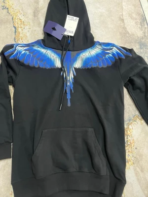 Svart hoodie med blå vingar - Säljer en svart hoodie från Marcelo Burlon med ett fett blått och vitt vingmönster över axlar och bröst. Hoodien har huva med dragsko och en stor magficka. Perfekt för dig som vill sticka ut med en unik design.