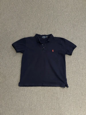 Ralph lauren piké - Klassisk marinblå pikétröja från Polo Ralph Lauren med röd broderad logga på bröstet. Slim fit-modell med korta ärmar och krage med knappar. Tillverkad i stretchig bomull som ger en bekväm passform.