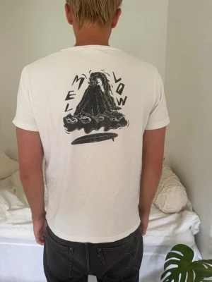 Vit t-shirt med tryck och surfbräda - Vit t-shirt i bomull med svart tryck på ryggen som föreställer en vulkan och texten 'MELLOW', samt en liten surfbräda på bröstet. Klassisk passform och korta ärmar, perfekt för en avslappnad stil. Köpt i Sri lanka 