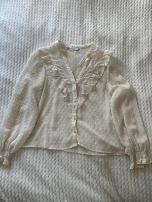 Vit blus med volang från Zara - Söt vit blus från Zara med volangdetaljer längs v-ringningen och knappar framtill. Blusen har långärmade ärmar med resår vid ärmslut och ett diskret prickigt mönster i tyget. Perfekt för en trendig och romantisk look.