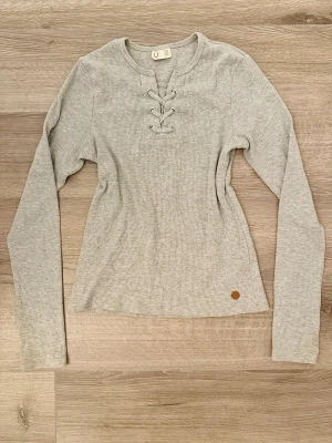 Ljusgrå tröja Kappahl 158/164 - Ljusgrå långärmad tröja i ribbad bomullsmix med snygg snörning vid halsen. Tröjan har en rundad halsringning och en liten patchdetalj nertill. Perfekt för en avslappnad och trendig look.