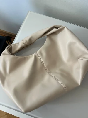 Beige axelremsväska i mjukt material - Trendig beige handväska i mjukt, slätt material med rundad form och bred axelrem. Väskan har synliga sömmar som ger en cool detalj och minimalistisk look. Perfekt för dig som gillar stilrena accessoarer med modern känsla.