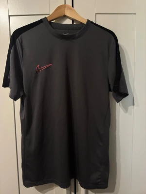 Mörkgrå Nike t-shirt med svarta detaljer - Snygg grå t-shirt från Nike med svarta paneler på axlarna och röd Nike-logga på bröstet. Klassisk rund hals och korta ärmar. Perfekt för en sportig och avslappnad stil. Den är slim fit och är L men passar L/M