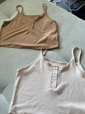 Två ribbade linnetoppar i beige toner - Säljer två ribbade linnetoppar med smala axelband och knappdetaljer framtill. Ena toppen är ljusbeige och den andra är i en mörkare beige nyans. Perfekta för sommaren och superenkla att matcha. Dem är som M
