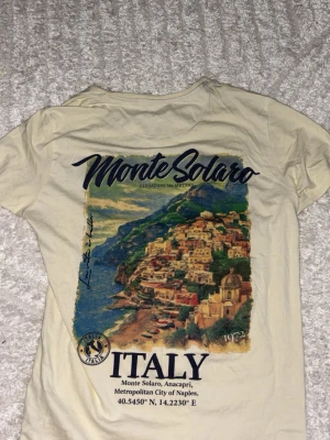 Beige Monte Solaro t-shirt med tryck - Snygg beige t-shirt med v-ringning och stort färgglatt tryck av Monte Solaro och italiensk kust på ryggen. Framtill finns en mindre logga med texten 'Monte Solaro Italy'. Perfekt för dig som gillar resor och coola motiv.