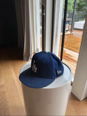 Blå LA New Era keps - Snygg mörkblå keps från New Era med LA-logga framtill i vitt och broderad New Era-logga på sidan. Klassisk basebollmodell med böjd skärm och broderade ventilationshål. Passar perfekt till streetwear och sportiga outfits.