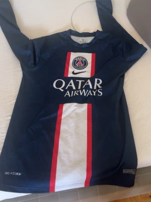 PSG långärmad matchtröja blå - Säljer en långärmad Paris Saint-Germain matchtröja i mörkblått med röd och vit rand längs mitten. Tröjan har tryck med Qatar Airways och Nike-logga på bröstet. Perfekt för fotbollsträning eller att bära som supporter.