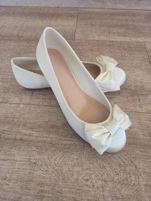 Satinskor i ballerinamodell  med rosett st 36 - Satinskor i ballerinamodell med rund tå och  dekorativ rosett framtill. Skorna har en klassisk och stilren design med låg klack och slät yta. Färg Ivory  i  lyxig satin