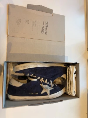 Marinblå Golden Goose- Superstar sneakers - Snygga blå sneakers från Golden Goose i använt, men bra och välbehållet skick. Köpta från Miinto för 5000 år 2024. Orginalkartong, äkthetsbevis-Golden Goose passport- och dustbag medföljer. Storlek 41-42 EU. 