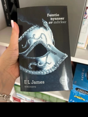Femtio nyanser av mörker pocket - En svensk pocketbok av E L James med titeln 'Femtio nyanser av mörker'. Omslaget är svart med en silverfärgad mask i fokus. Boken ges ut av Norstedts och är en del av den populära Fifty Shades-serien.