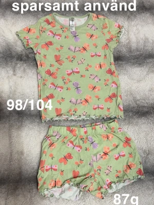 Grön pyjamas med fjärilar och blommor - Söt tvådelad pyjamas i ljusgrönt med färgglada fjärilar och blommor. Setet består av en kortärmad topp och shorts med volangkant. perfekt för mysiga kvällar.🌸 Sparsamt använd. Storlek 98/104