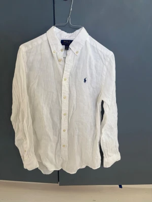 Vit linneskjorta från Polo Ralph Lauren - Klassisk vit skjorta i linne från Polo Ralph Lauren med button-down krage och broderad logga på bröstet. Storleken är 14/16 år 