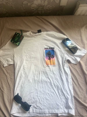 Vit t-shirt med Miami-tryck - Säljer en vit t-shirt med färgglatt Miami Florida-tryck framtill. Motivet visar palmer mot en solnedgång i blått, rosa och gult. T-shirten har klassisk passform och korta ärmar. Perfekt för dig som gillar streetstyle och somriga vibes. Pris kan diskuteras.