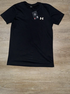 Svart t-shirt från Under Armour - Svart t-shirt från Under Armour med klassisk logga på bröstet. T-shirten har rund halsringning och korta ärmar. Perfekt för en sportig och avslappnad stil.