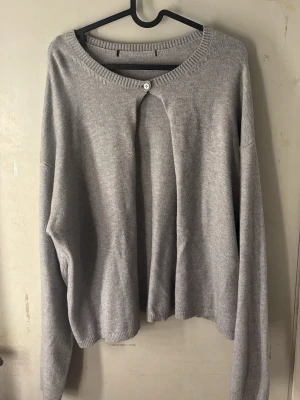 Ljusgrå kofta med knapp - Ljusgrå, tunnstickad kofta med rund halsringning och en knapp framtill. Koftan har långa ärmar och en avslappnad passform. Perfekt att slänga över en topp för en enkel och stilren look.