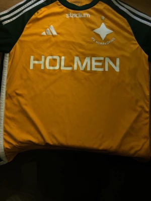IFK Norrköping matchtröja Adidas gul - Gul IFK Norrköping matchtröja från Adidas med gröna detaljer och vita ränder på axlarna. Tryck med 'HOLMEN' på bröstet och klubbmärke. Tillverkad i lätt och funktionellt material, perfekt för träning eller match.