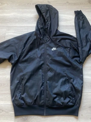 Svart vindjacka med huva från Nike - Snygg svart vindjacka från Nike med dragkedja framtill, två fickor med dragkedja och justerbar huva. Jackan har en sportig look med Nike-logga på bröstet och är tillverkad i ett lätt, glansigt material som står emot vind. Mycket bra skick använd fåtal gånger. 