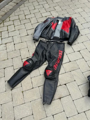 Dainese svart och röd mc-skinnställ - Säljer ett tvådelat mc-skinnställ från Dainese i svart och rött. Jackan har röda detaljer och logga på ärmen, byxorna har stora röda Dainese-tryck och knäskydd. Tillverkat i slitstarkt läder