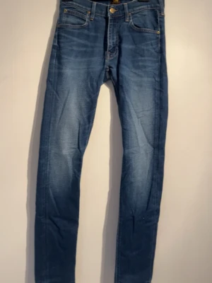 Blå slim jeans från Lee - Säljer ett par blå slim jeans från Lee med klassisk femficksmodell och guldfärgade detaljer. Jeansen har en snygg tvätt med lätt slitning och är tillverkade i stretchigt denimtyg för skön passform. Perfekta för dig som gillar en smal siluett.