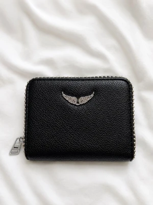 Svart plånbok från Zadig & Voltaire - Snygg svart plånbok i läder från Zadig & Voltaire med silverfärgad dragkedja och ikoniska vingloggan framtill. Plånboken har flera kortfack, myntfack med dragkedja och detaljer i silvermetall. Perfekt storlek för att få plats med allt viktigt.