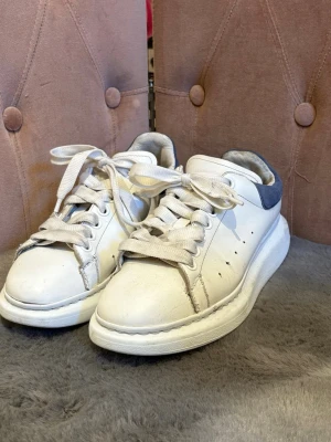 Alexander McQueen vita sneakers - Stilrena vita sneakers från Alexander McQueen med bred sula och blå mockadetalj på hälen. Klassisk snörning och diskret logga baktill. Skorna är tillverkade i läder med detaljer i mocka och har en chunky siluett som ger en modern look.