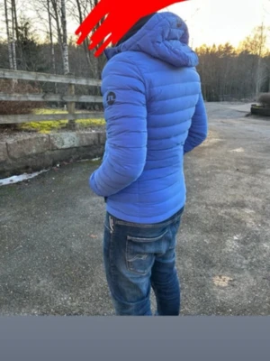 Blå pufferjacka från J.Lindeberg - Snygg blå pufferjacka från J.Lindeberg med huva och dragkedja framtill. Jackan har ett quiltat mönster och logotyp på ärmen. Perfekt för kalla vinterdagar och ger en sportig look.
