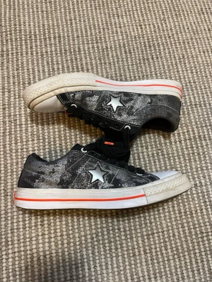 Converse One Star X Sad Boys - Svarta Converse One Star sneakers med vit stjärna på sidan, svartvitt mönster och röda detaljer. Skorna har vita sulor med röd rand, svarta skosnören och grafiskt rödblått innerfoder. Klassisk låg modell i canvasmaterial.