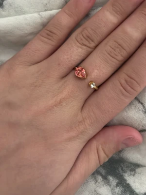 Caroline svedbom - Trendig guldfärgad ring med öppen design, prydd med en stor rosa sten och en mindre vit sten. Ringen har en tvinnad detalj på bandet som ger en cool och modern känsla. Perfekt accessoar för att lyfta din stil.