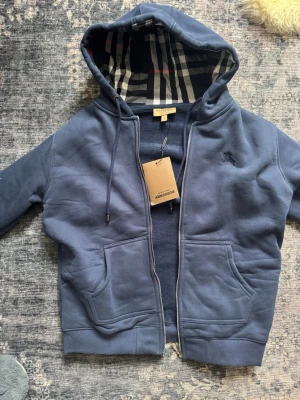 Blå hoodie från Burberry med dragkedja - Snygg blå hoodie från Burberry med dragkedja och klassiskt rutigt foder i huvan. Hoodien har två stora fickor framtill, dragsko i huvan och broderad Burberry-logga på bröstet. Perfekt för en chill och stilren look.