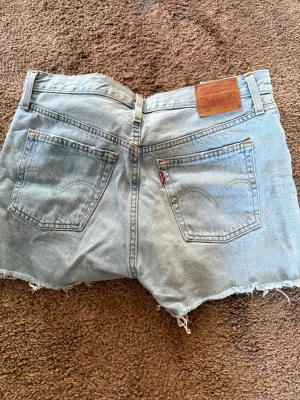 Levi's 501 ljusblå jeansshorts - Ljusblå jeansshorts från Levi's modell 501 med klassisk femficksdesign, råa kanter och läderpatch bak i midjan. Shortsen har en avslappnad passform och är tillverkade i bomull. Perfekta för sommardagar och enkel att matcha med allt.